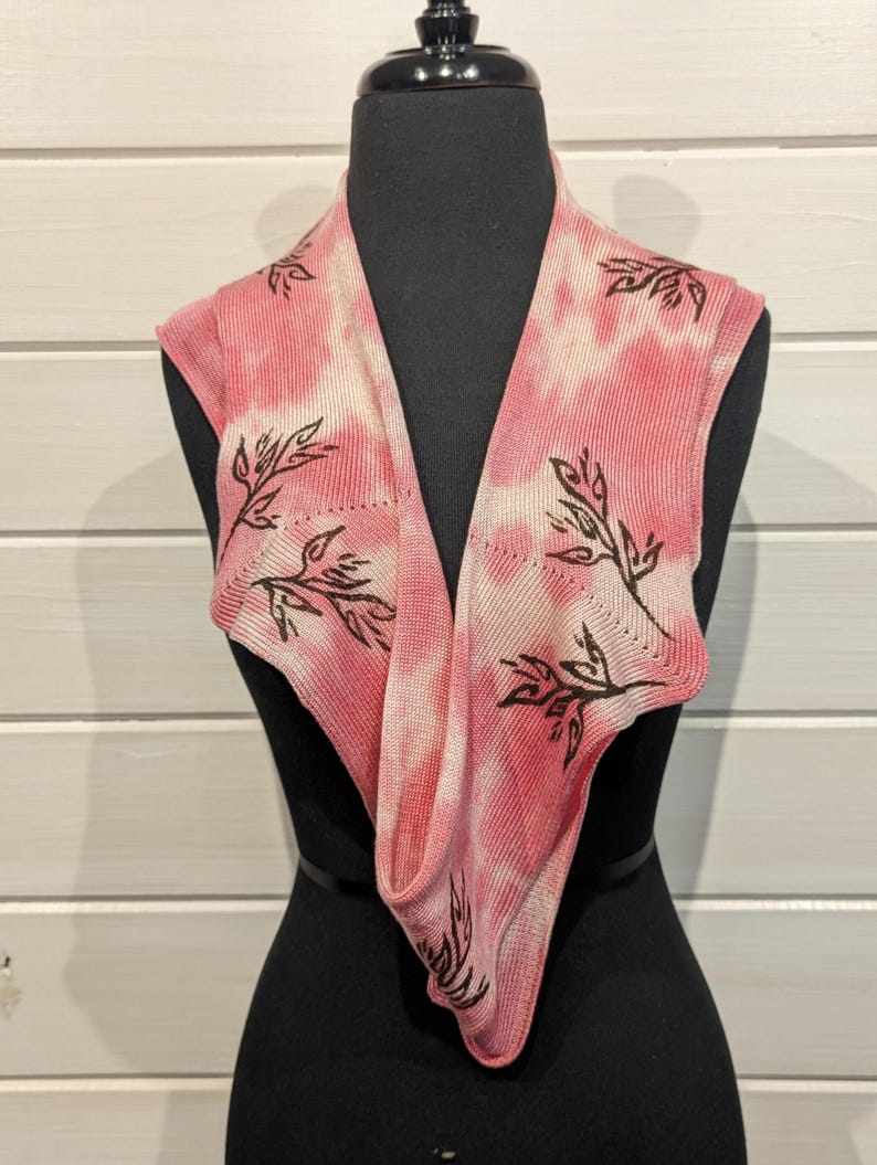 May include: A pink and white tie-dye knit cowl scarf with a leaf design. The scarf is displayed on a black mannequin. The leaf design is in a dark brown color. The scarf is a soft, lightweight accessory.