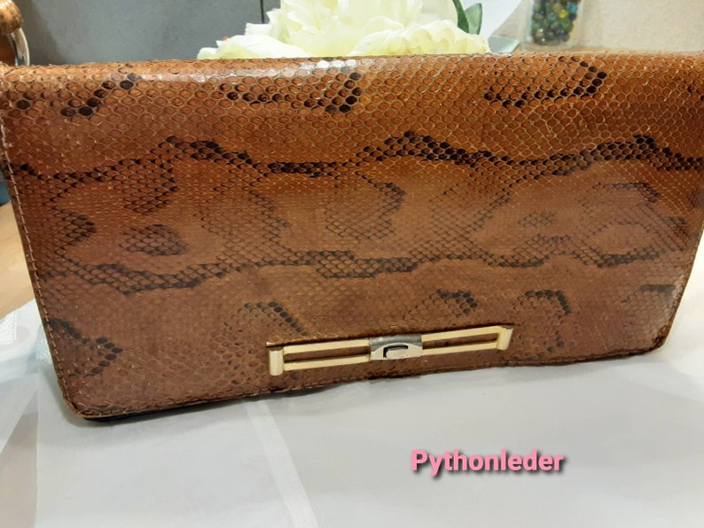 May include: A brown leather clutch bag with a snakeskin pattern and a gold metal clasp. The bag has the text "Pythonleder" printed on the bottom.