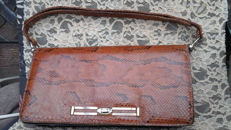 May include: A brown snakeskin clutch bag with a silver metal clasp. The bag has a strap and is made of genuine leather.