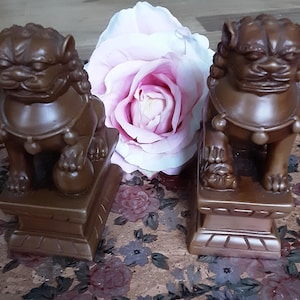 Könnte beinhalten: Zwei braune Foo-Dog-Statuen mit detaillierten Merkmalen, die jeweils eine Kugel halten, sitzen auf einem dekorativen Sockel. Eine große rosa-weiße Rose ist zwischen ihnen positioniert. Die Statuen haben eine warme, braune Farbe.