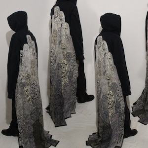 May include: Black hooded sweatshirt with a long, flowing back panel featuring a detailed, grayscale illustration of skulls and figures. The design has a gradient effect, transitioning from light to dark.