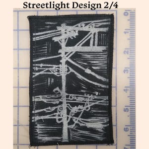 May include: A black and white embroidered patch with a design of a streetlight and power lines. The patch is rectangular and has a white border. The text "Streetlight Design 2/4" is printed above the patch.