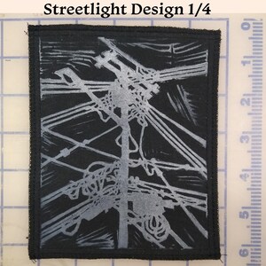 Op de afbeelding: Een zwarte rechthoekige patch met een zilveren ontwerp van een straatlantaarn en elektriciteitsleidingen. De tekst "Streetlight Design 1/4" staat bovenaan. De patch heeft een zwarte rand en is waarschijnlijk een strijk- of opnaaipatch.