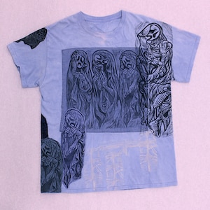 Op de afbeelding: Lichtblauw T-shirt met een donkerblauwe grafische print. De afbeelding toont gestileerde figuren met expressieve gezichten en vloeiende gewaden. Extra donkerblauwe grafische elementen zijn geplaatst op de mouwen en de onderkant van de voorkant. Het T-shirt heeft een ronde hals.