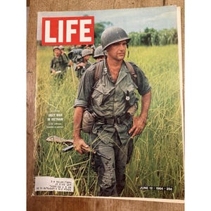 May include: A vintage LIFE magazine cover from June 12, 1964, featuring a U.S. officer in Vietnam. The cover has the headline "Ugly War in Vietnam" in white text. The officer is wearing a helmet and military uniform, walking through tall grass.