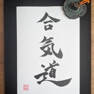 Könnte beinhalten: Ein weißes rechteckiges Papier mit schwarzer japanischer Kalligrafie für Aikido, montiert auf einem schwarzen Rand. Ein kleines, dunkles, blumenförmiges Metallobjekt mit einer orangefarbenen Kordel befindet sich oben rechts und ein rotes quadratisches Siegel unten links.