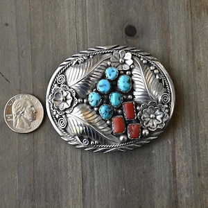 May include: Oval silver belt buckle with turquoise and coral stones. The buckle features a floral and leaf design with a central cluster of turquoise stones and rectangular coral stones. A quarter coin is next to the buckle for size comparison.