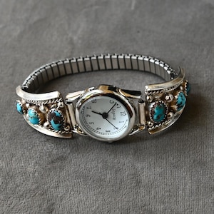 Op de afbeelding: Zilverkleurig horloge met een witte wijzerplaat en zwarte wijzers. De horlogeband is een rekbare stijl. Het horloge is versierd met turquoise gekleurde stenen gezet in decoratieve zilveren zettingen. Het horloge ligt op een grijze ondergrond.