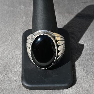 Peut inclure: Une bague en argent avec une pierre noire ovale. La bague présente un motif détaillé autour de la pierre. Elle est présentée sur un support noir sur fond gris. La bague est un bijou.