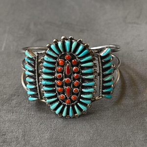 Op de afbeelding: Zilveren manchetarmband met een ovaal middelpunt van turkoois en koraal. Druppelvormige turkooizen stenen omringen een centrale cluster van koraalstenen. De armband heeft een ontwerp met drie banden en decoratieve zij-elementen.