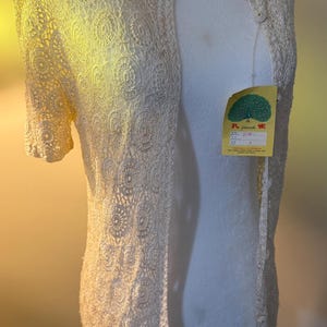 May include: Cream-colored, open-front cardigan with short sleeves. The intricate lace design features a repeating circular pattern. The garment is displayed on a mannequin, with a tag attached. The tag has Japanese text and a peacock illustration.