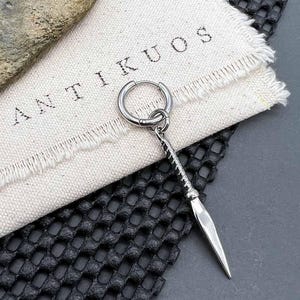 May include: A silver hoop earring with a silver dagger pendant. The earring is on a white fabric surface with the word "ANTIKUOS" printed on it.