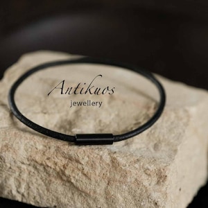 Peut inclure: Un bracelet en cordon noir fin doté d'un fermoir cylindrique en métal noir lisse. Le texte Antikuos jewellery est imprimé en noir, en écriture cursive et en police sans empattement, au centre du cordon circulaire posé sur une pierre beige brute.