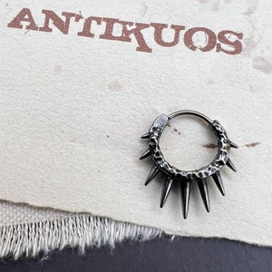 May include: A black metal hoop earring with a textured surface and six sharp points extending from the hoop. The earring is on a white background with the word "ANTIKUOS" printed in brown.