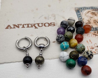 May include: A pair of silver hoop earrings with a black and a gold bead dangling from each hoop. The earrings are displayed on a white surface with a variety of colorful stones.