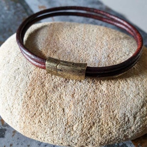Pode incluir: Uma pulseira de couro castanho com fecho magnético. A pulseira é feita de duas tiras de couro e tem um fecho de cor bronze. A pulseira está sobre uma pedra castanha clara.