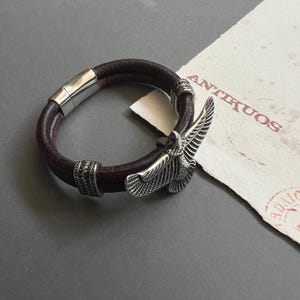 以下が含まれることがあります： 銀色の鷲のチャームが付いた茶色の革のブレスレット。ブレスレットは磁気留め金で留められ、小さな銀色のビーズで飾られています。