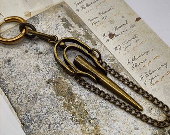 Può includere: Una catena dorata con una chiusura decorativa e una punta appuntita. La catena è attaccata a un orecchino a cerchio. La catena è su uno sfondo bianco con testo scritto a mano.