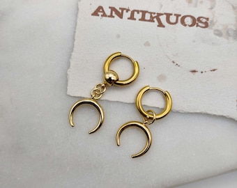 May include: A pair of gold hoop earrings with crescent moon charms hanging from them. The earrings are on a white surface with the word "ANTIKUOS" printed on a piece of paper behind them.