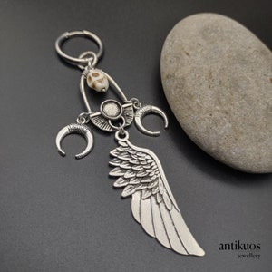 Può includere: Ciondolo in argento con un'ala, due mezze lune, un ciondolo a forma di teschio e una perla rotonda. Il ciondolo è attaccato a un anello d'argento.