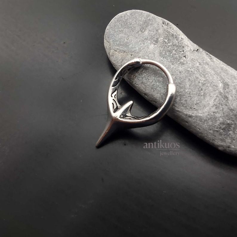 Puede incluir: Pendiente de aro de plata con un dise&ntilde;o puntiagudo detallado. El pendiente est&aacute; sobre un fondo de piedra gris. El texto "antikuos jewellery" es visible debajo del pendiente.