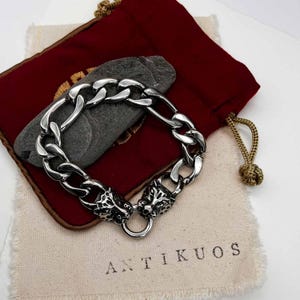 May include: A silver chain bracelet with a chunky link design. The bracelet has two detailed lion head clasps. The bracelet is displayed on a stone, a red velvet pouch, and a canvas bag with the text "ANTIKUOS".