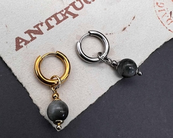 May include: Two hoop earrings, one gold-toned and one silver-toned, each with a gray stone bead pendant. The earrings are displayed on a textured paper with the word "ANTIKUOS" stamped on it. The background is dark.