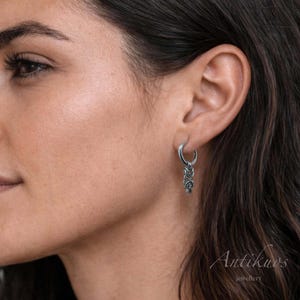 May include: A silver hoop earring with a dangling, ornate design. The earring is attached to the ear. The design is made of several interconnected, textured elements. The word "Antikuos" and the word "jewellery" are visible in the bottom right corner.