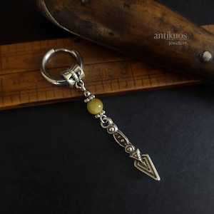 May include: A silver hoop earring with a silver charm hanging from it. The charm is a small arrow with a tiger's eye bead above it. The earring is on a wooden ruler.