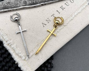 May include: Two sword-shaped earrings, one silver and one gold, displayed on a white cloth with the word "ANTIKUOS" printed on it. The earrings have a hoop closure. A large, textured metal object is in the background.