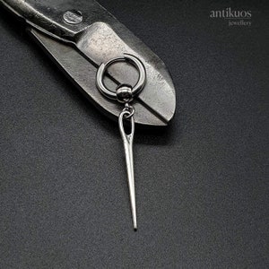 May include: A silver earring with a needle pendant hangs from a hoop. The earring is displayed on a pair of metal shears against a dark background. The earring has a small ball at the top of the needle.
