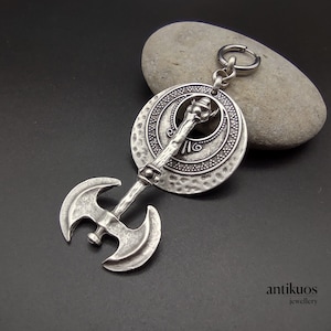 May include: Silver double-headed axe pendant with a round, textured disc featuring a circular design. The pendant is attached to a silver chain with a silver ring.