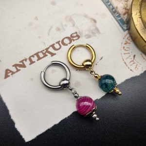 Könnte beinhalten: Zwei Creolen mit baumelnden Edelsteinperlen. Die Creole links ist silberfarben und hat eine rosafarbene Achatperle. Die Creole rechts ist golden und hat eine grüne Achatperle.