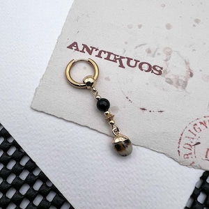 Puede incluir: Pendiente de aro de oro con una cuenta negra y un dije de piedra moteada.