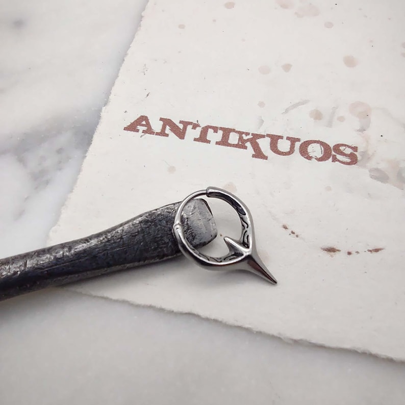 Puede incluir: Un aro de plata para la oreja con un dise&ntilde;o puntiagudo. El aro est&aacute; sobre una superficie blanca con la palabra "ANTIKUOS" impresa en marr&oacute;n.