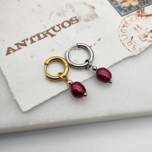May include: Two hoop earrings with a dangling burgundy pearl. One earring is gold and the other is silver.