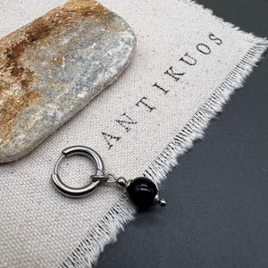 Peut inclure: Une boucle d'oreille en argent avec un charme en perle noire suspendu. La boucle d'oreille est sur un fond en tissu blanc avec le mot "ANTIKUOS" imprimé dessus.