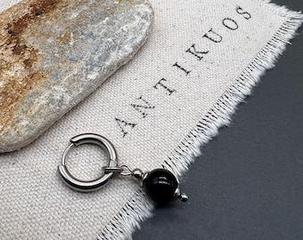 May include: A silver hoop earring with a black bead charm hanging from it. The earring is on a white fabric background with the word "ANTIKUOS" printed on it.