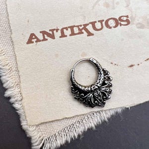 以下が含まれることがあります： 花柄の細工が施されたシルバーのフープピアス。ピアスは、"ANTIKUOS"という文字が印刷された白い背景の上に置かれています。