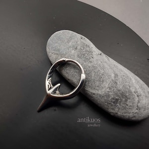 Puede incluir: Pendiente de aro de plata con un dise&ntilde;o detallado y una punta puntiaguda. El pendiente est&aacute; sobre una superficie de piedra gris.