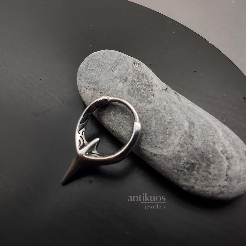 Puede incluir: Pendiente de aro de plata con un dise&ntilde;o detallado y una punta puntiaguda. El pendiente est&aacute; sobre una superficie de piedra gris.