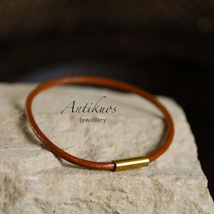 Pode incluir: Uma pulseira fina e redonda de cordão de couro castanho com um fecho cilíndrico de metal dourado polido. O texto Antikuos jewellery está impresso a preto no centro da pulseira circular, que está posicionada sobre uma superfície de pedra.