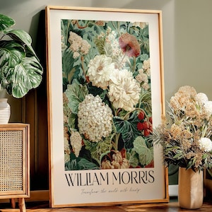 May include: A framed art print featuring a floral design with large white and cream-colored flowers, green leaves, and red berries. The print is labeled "WILLIAM MORRIS" with the text "Transform the world with beauty" below. The frame is a light brown color.