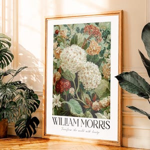 Op de afbeelding: Een ingelijste bloemenprint met een botanische illustratie van witte, rode en gele bloemen. De print is getiteld "WILLIAM MORRIS" met de tekst "Transform the world with beauty" eronder. De lijst is lichtbruin.