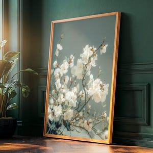 May include: A framed art print depicting white cherry blossom flowers against a muted teal backdrop. The artwork is in a light brown wooden frame, leaning against a dark green wall. A potted plant is visible in the foreground.