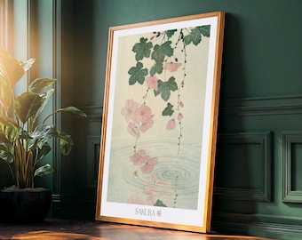 Op de afbeelding: Een ingelijste kunstdruk met een Japanse illustratie van roze kersenbloesems en groene bladeren boven water. De print heeft een lichtbeige achtergrond en is ingelijst in een licht houten frame. Het woord "SAKURA" staat onderaan afgedrukt.