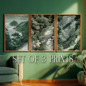 May include: A set of three framed prints displaying landscape art. The prints depict mountainous scenery with green trees, a river, and clouds. The text "SET OF 3 PRINTS" is visible below the framed pictures. The frames are a light brown color.