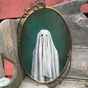 May include: A white ghost painted on a canvas in an oval, ornate, gold-colored frame. The ghost is wearing a white sheet and has two black eyes.