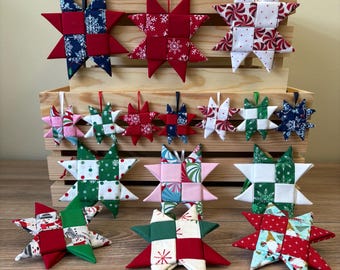 May include: A collection of fabric star ornaments in various patterns and colors, including red, green, white, blue, and pink. Each star features a quilted, multi-paneled design with a hanging loop, displayed in a tiered wooden crate arrangement.