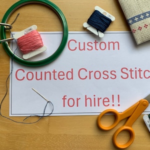 May include: A white card with the text "Custom Counted Cross Stitcher for hire!" in red letters.  The card is surrounded by embroidery supplies, including a green embroidery hoop, a needle, blue and pink embroidery floss, and a pair of orange scissors.